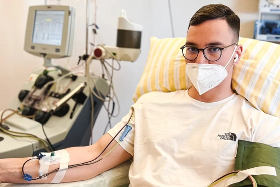 Stammzellspender Bastian sitzt auf dem Krankenbett. Im Hintergrund ist die Zentrifuge für Stammzellentnahme zu sehen.