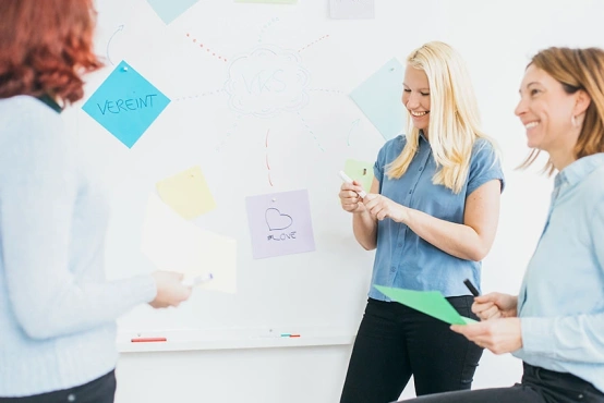 Annett, Katja und Juliane vom VKS Team stehen vor einer Brainstorming Wand, mit Zettel und Stiften in der Hand, um neue Pläne und Ideen zu notieren.