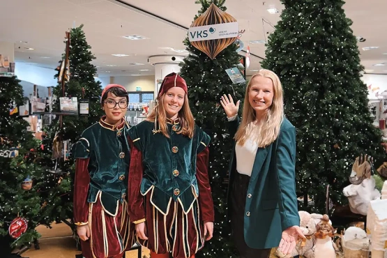 Drei fröhliche Personen im Charity-Wald der Galeria Kaufhof in Chemnitz, darunter Katja in einem eleganten grünen Blazer, flankiert von zwei lächelnden Helfern in traditionellen Weihnachtselfenkostümen, die die festliche Spendenaktion unterstützen