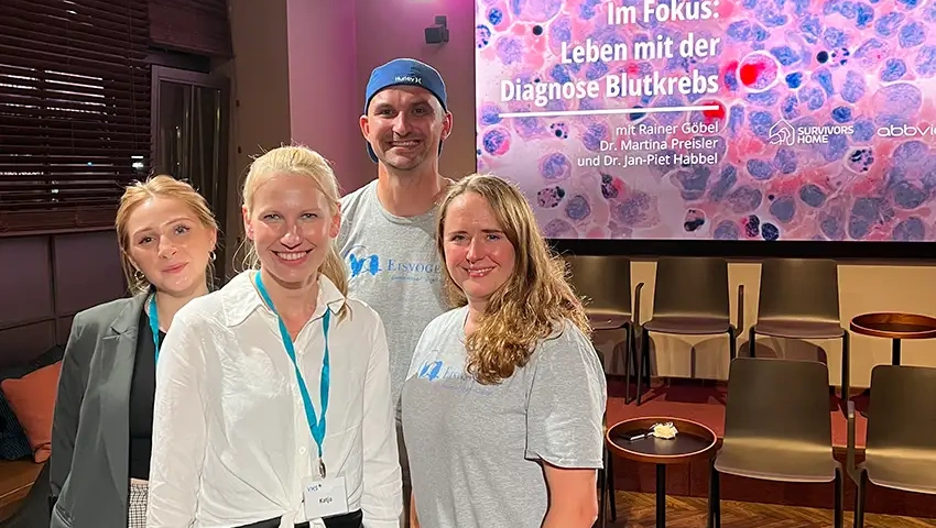 Engagierte Mitglieder von Survivors Home und dem VKS posieren lächelnd vor einem informativen Display über Leukämie beim Survivors Home Event in Berlin, das Aufmerksamkeit und Unterstützung für das Leben mit der Diagnose Blutkrebs fördert