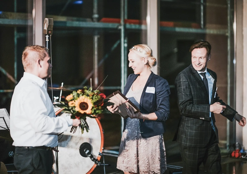 Zwei VKS Vorstandsmitglieder und Moderatorin Henriette Fee stehen auf der Bühne vom Stammzellspenderempfang 2019. Dabei wird Henriette einen Blumenstrauß überreicht, dabei strahlen alle.