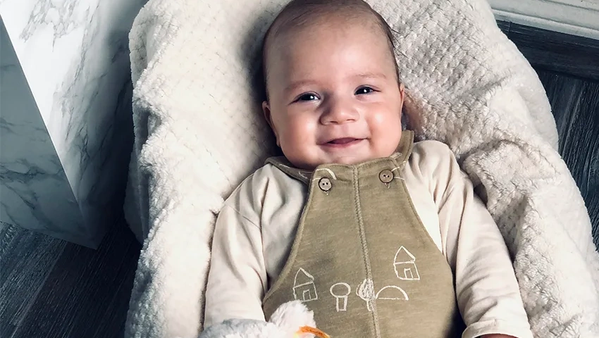 Ein strahlendes Baby namens Mahir, gekleidet in einem niedlichen, olivgrünen Strampler mit Spielzeughase, lächelt fröhlich, nachdem er eine lebensrettende Stammzelltransplantation erhalten hat
