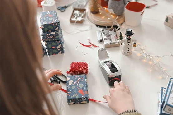 Eine Person verpackt Weihnachtsgeschenke mit Bastelpapier und festlichen Dekorationen auf einem Tisch und zeigt den intimen Prozess des Personalisierens von Feiertagsgeschenken.