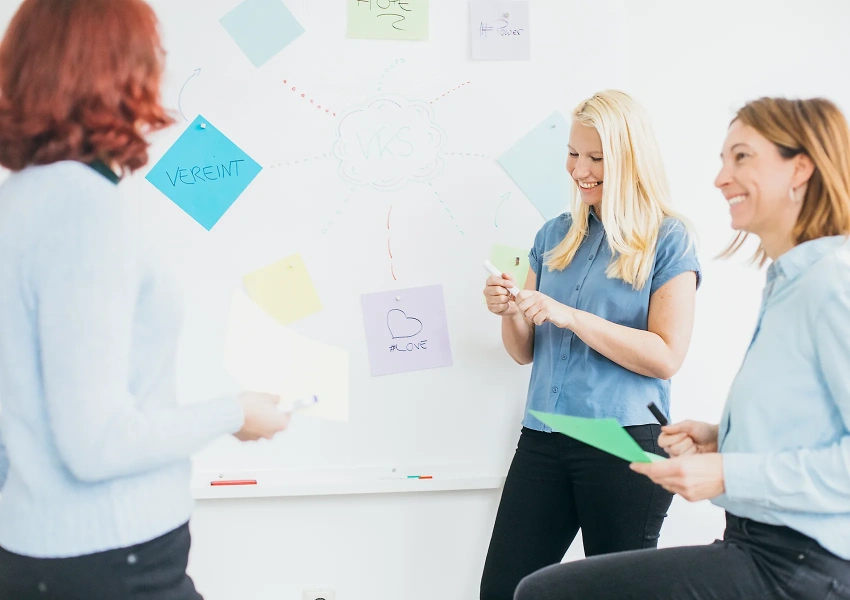 Annett, Katja und Juliane vom VKS Team stehen vor einer Brainstorming Wand, mit Zettel und Stiften in der Hand, um neue Pläne und Ideen zu notieren.