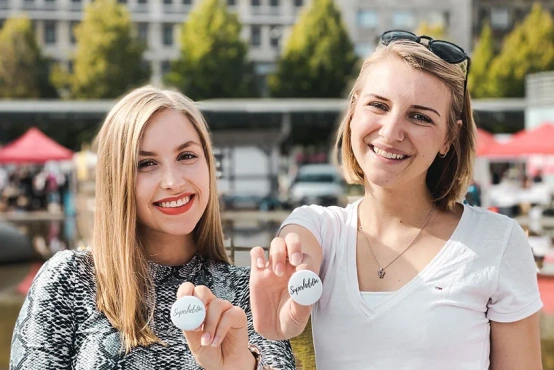 Zwei potenzielle Spenderinnen auf einer VKS Typisierungsaktion lächeln und halten einen Superhelden Button nach vorn. 