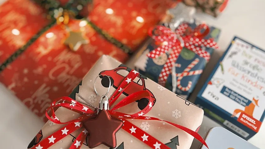 Verschiedene Weihnachtsgeschenke im Closeup in buntem Papier mit Bändern und Verzierungen verpackt, einschließlich Bücher 'Stadt Land Fluss' und 'Der neue große Wissenstest für Kinder', die den Geist der Saison vermitteln.