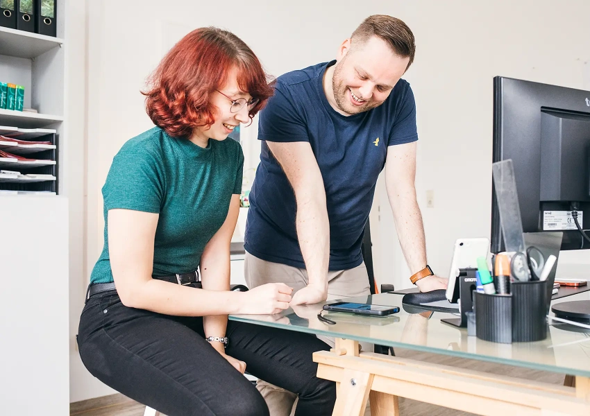 Sebastian und Annet vom VKS Team sitzen gemeinsam am Arbeitsplatz und schauen auf das Handy. Sie planen zusammen die nächste Social Media Story über einen Stammzellspender.