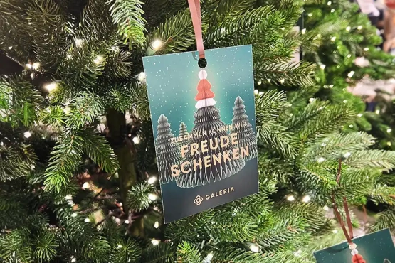 Eine dekorative Weihnachtsbaumkarte hängt zwischen üppig grünen Zweigen, beschriftet mit 'Zusammen können wir Freude schenken'.